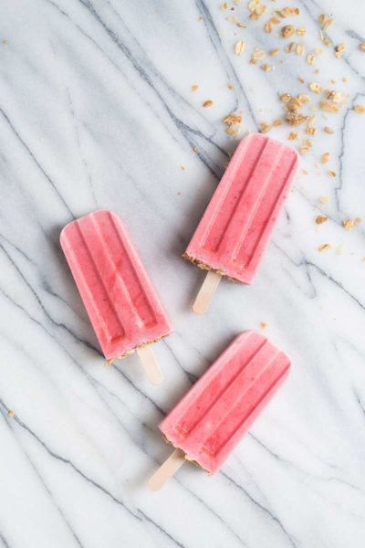 overhead image: 3 strawberry yogurt popsicles with granola on gray marble background
