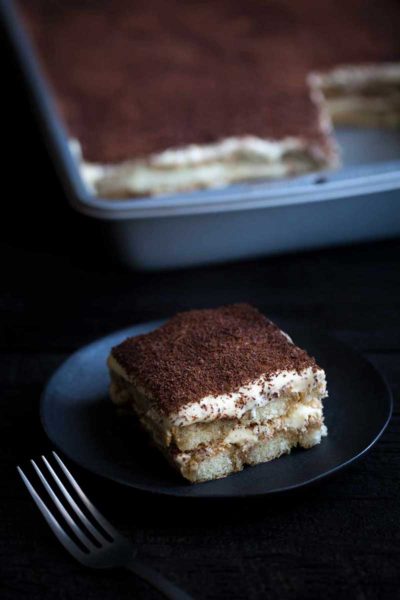 Closeup of tiramisu serving on a plate with full pan of tiramisu in the background