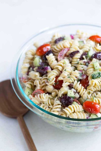 A close up shot of Greek pasta salad in a bowl, next to a wood spoon.