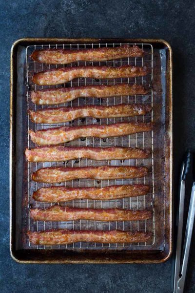 Oven-baked bacon on a sheet pan and wire rack. Photo shows overhead shot of finished bacon on the sheet pan and tongs on a blue backdrop.