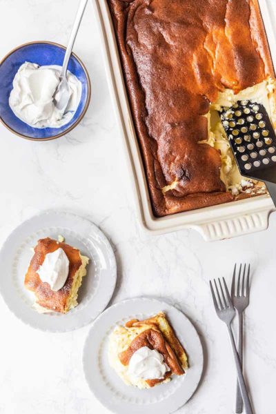 Two servings of blintz souffle on plates, topped with sour cream and sugar, sitting next to the remaining souffle